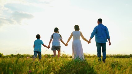 Fototapeta premium Happy family walks through green meadow, unity, support, cohesion, slow motion. Parents father mother, children daughter son hold hands, family picnic in nature. People enjoy freedom of outdoors, sky