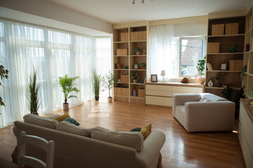 Bright modern living room with large windows, cozy furniture, wooden flooring and indoor plants creating a warm, inviting interior.
