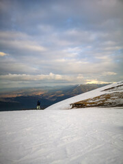 Person with snowboard standing on snowy mountain slope looking at wide winter landscape with soft clouds and warm sunset tones in vertical format.