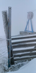 Cold winter scene with frosted metal fence and ski lift machinery in dense fog, depicting harsh mountain weather and minimal ambience.