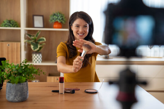 Smiling young woman presenting a makeup brush while recording beauty content for social media in a home studio.

