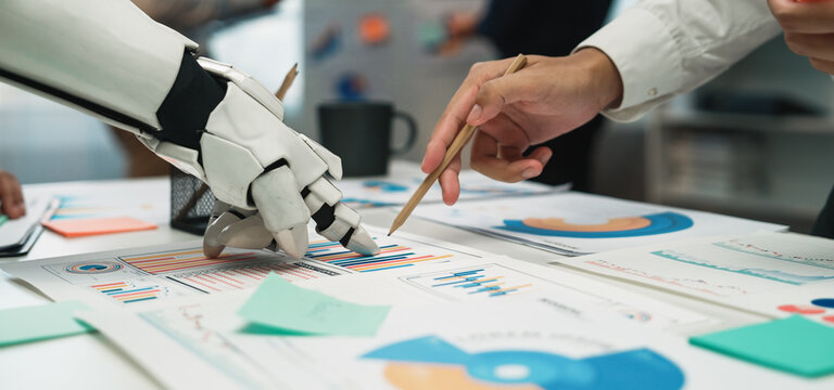 A modern office scene showcasing collaboration between a robotic hand and a human hand, analyzing various data charts and graphs for innovative business solutions.Noogenesis