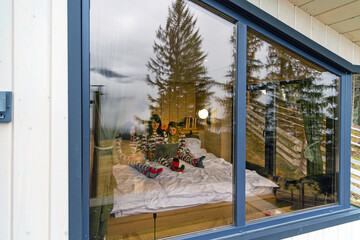 Family in Christmas pajamas seen through cabin window. View from outside a cozy wooden cabin showing mother and daughters in festive pajamas watching a laptop together on a bed.