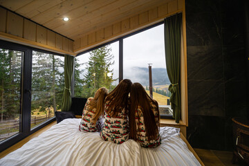 Family in matching pajamas admiring Mountain View. Three girls in festive Christmas pajamas sitting on a bed, peacefully looking out large cabin windows at the scenic mountain landscape.