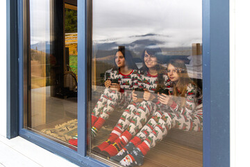 Family in pajamas enjoying cabin view indoors. Three girls wearing festive pajamas sit inside a cozy cabin, holding mugs and looking out at a scenic mountain landscape.