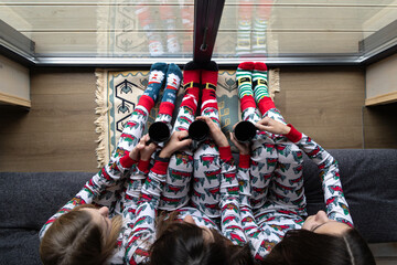 Top view of family in pajamas with mugs. Three girls wearing festive pajamas and colorful socks hold mugs while relaxing indoors, seen from above near a glass window.