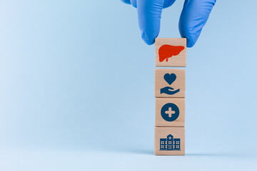 A doctor wearing a blue glove sorts wooden cubes with a liver icon and medical and health icons on a light blue background. Concept of liver donation and organ donation awareness.