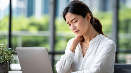 Overworked Asian office employee suffering from neck and shoulder pain, massaging her neck while sitting in front of a laptop. Stressful work environment, poor posture, and long sc