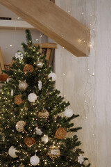 Decorated Christmas tree with warm lights and rustic ornaments stands in a loft room with a wooden beam and light textured wall. Suitable for holiday themes, winter decor, celebration concepts.