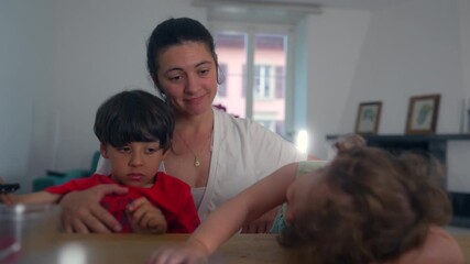 Mother sitting with children at table while toddler leans forward with lively curiosity and older child remains calm, forming layered family moment rich in motion and gentle closeness