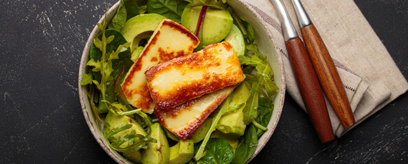 Fried halloumi cheese and sliced avocado salad with mixed green leaves, healthy and gourmet vegetarian meal, black background