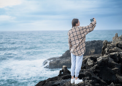 Caucasian Woman Taking Selfie By The Atlantic Ocean On A Rocky Shore. Portugal - Powered by Adobe