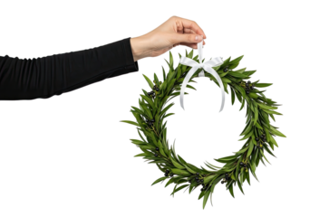 A woman's hand in a black sleeve holds a beautiful festive wreath made of fresh green leaves and dark berries, tied with a white satin bow and isolated on a black background