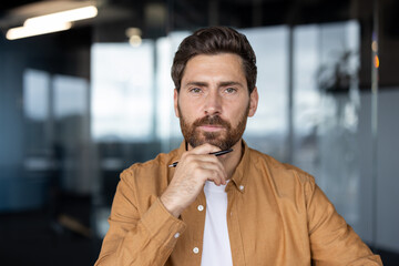 Naklejka premium Serious well-groomed professional man having an idea, looking at camera while holding a pen under his chin, planning strategy in a contemporary workplace environment