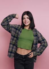 portrait of confident young woman poses with one arm raised, showcasing her strength and style in a green top and plaid shirt on pink background