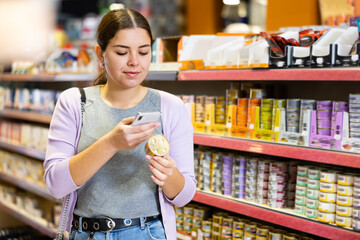 Positive young female customer scanning barcode on tin can of canned food for cats with smartphone while shopping in pet store, paying for item using mobile app..
