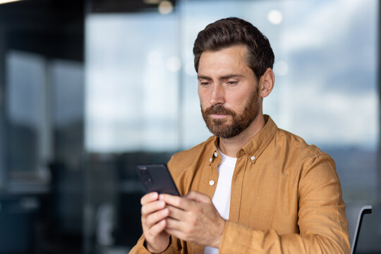 Bearded man sitting in modern office interior, focused on typing message or browsing content on a mobile phone, representing technology use, digital communication, and online business activity