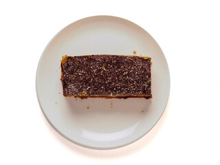 A dessert layered cake with chocolate is on a white plate on white background. The dessert is a cake with a brown crust. The plate is white and the dessert is the main focus of the image