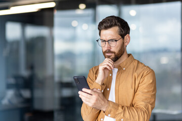 Man wearing glasses closely examining a smartphone screen while concentrating on complex data, solving a problem, or receiving unexpected news in a modern office environment