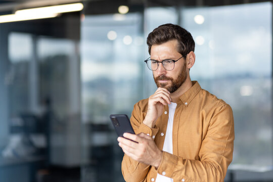 Man wearing glasses closely examining a smartphone screen while concentrating on complex data, solving a problem, or receiving unexpected news in a modern office environment - Powered by Adobe