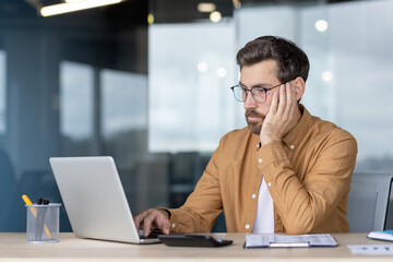 Businessman exhausted and bored at a desk, leaning on hand while typing on a laptop in a modern home office, showing stress, burnout and deadline pressure