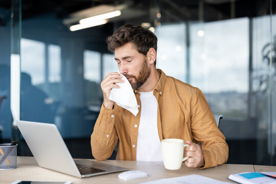 Man with a beard holding a tissue and blowing his nose while sitting at his desk, working on a laptop in a modern office, experiencing cold or flu symptoms