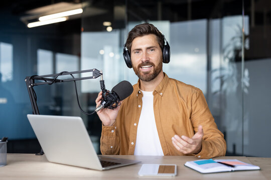 Man wearing headphones and orange shirt broadcasting, talking into a professional microphone at a desk with a laptop and notebook, creating engaging audio content - Powered by Adobe