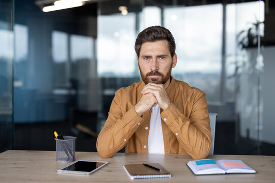 Thoughtful bearded man in a brown shirt looking directly at the camera while sitting at his office desk, reflecting on business ideas, challenges, and growth opportunities