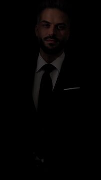 Attractive man in a suit is looking down, smiling, looking away, and then fixing his tie on dark studio background.