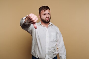 portrait of young man displays a thumbs down gesture with a frown, showcasing disapproval on beige background