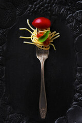 A fork with pasta, tomato sauce, a basil leaf and a cherry tomato on a black serving platter.
