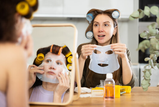 Two female friends sharing moment of relaxation and enjoying spa day at home, sitting by tabletop vanity mirror with colorful rollers in hair and applying moisturizing sheet masks to faces