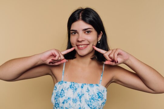 portrait of young woman smiles while pointing to her cheeks, showcasing a friendly and approachable vibe on beige background