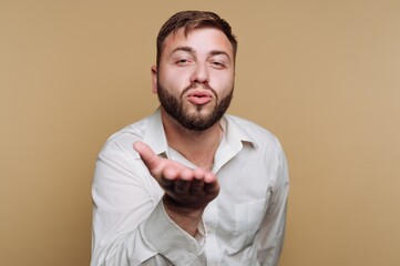 portrait of charming young man blows a kiss towards the camera, reflecting warmth and affection on beige background