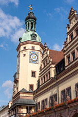 Historic tower ornate clock rises above vibrant Leipzig architecture under clear blue sky. Warm sunlight highlights detailed facade, creating vivid contrast soft clouds elegant decorative elements