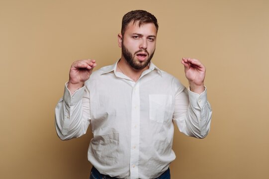 portrait of young man gestures widely with his hands while exhibiting a puzzled expression on beige background