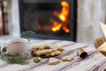 Christmas gingerbread cookies, warm cup of tea, fir branches and ornaments on table on background of festive decorated burning fireplace. Merry Christmas! Cozy winter time