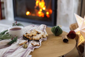 Merry Christmas! Gingerbread christmas cookies, warm cup of tea, fir branches and ornaments on table on background of festive decorated fireplace mantel with lights. Cozy winter time