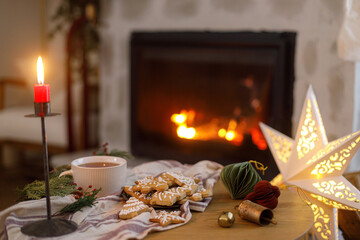 Merry Christmas! Gingerbread christmas cookies, warm cup of tea, fir branches and ornaments on table on background of festive decorated fireplace with lights in eve. Cozy winter time