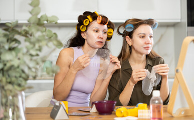 Two interested adult women testing out hydrating face paper mask during daily self-care routine, sharing thoughts on skincare experiences in cozy home environment