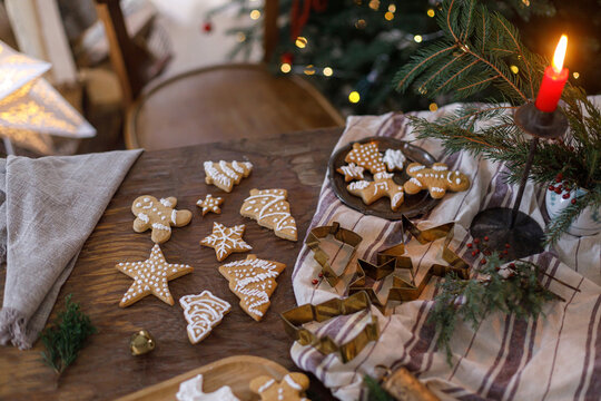 Gingerbread christmas cookies on rustic table with metal cutters, napkin, fir branches and candle on background of christmas tree lights. Christmas family traditions, holiday cookies