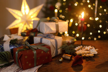 Merry Christmas! Stylish wrapped christmas presents, ornaments, candle, gingerbread cookies on table against festive decorated christmas tree with lights. Atmospheric holiday eve