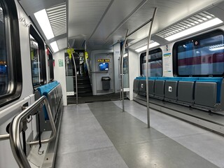 The interior of an empty, modern commuter or regional train carriage, featuring gray and blue seats, silver grab rails, and illuminated overhead lights. 