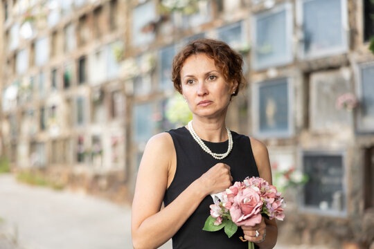 Sorrowful young woman in black dress with bouquet of flowers and handkerchief in hands, stands in cemetery. Visiting grave of deceased relative, mourning dead loved one. Appeal to God, prayer for dead - Powered by Adobe