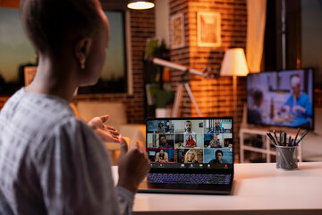 Young black woman working remotely from cozy living room during sunset, attending online video conference. Female entrepreneur having virtual discussion with diverse coworkers on laptop.