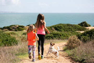 Two young ladies dressed in bright sport clothes are jogging with a dog on cliffs along the ocean 