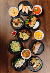 Assorted restaurant dishes flat lay on wooden table with soups salads mains bread and sauces