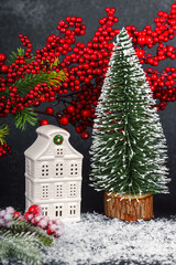 Ceramic Christmas village house with red holly berries, and small tree on black textured background with snow. Winter holiday home decoration, festive details, warm seasonal mood
