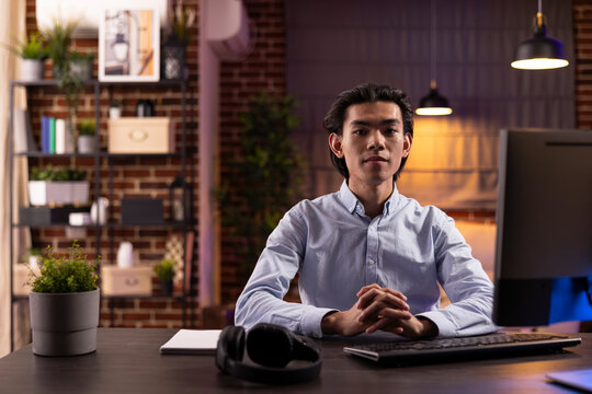 Serious young man sits at desk in home office ready to study online, research on internet and complete freelance tasks. Portrait of asian male entrepreneur with hands clasped, looking at camera.