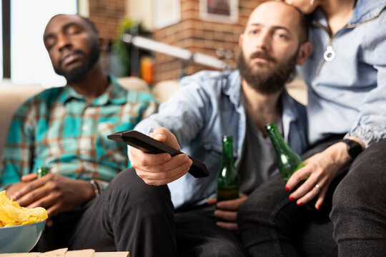 Close up of caucasian man using remote to switch channels on television. White bearded man adjusts volume and changes shows while friends sit nearby on couch during casual home hangout.